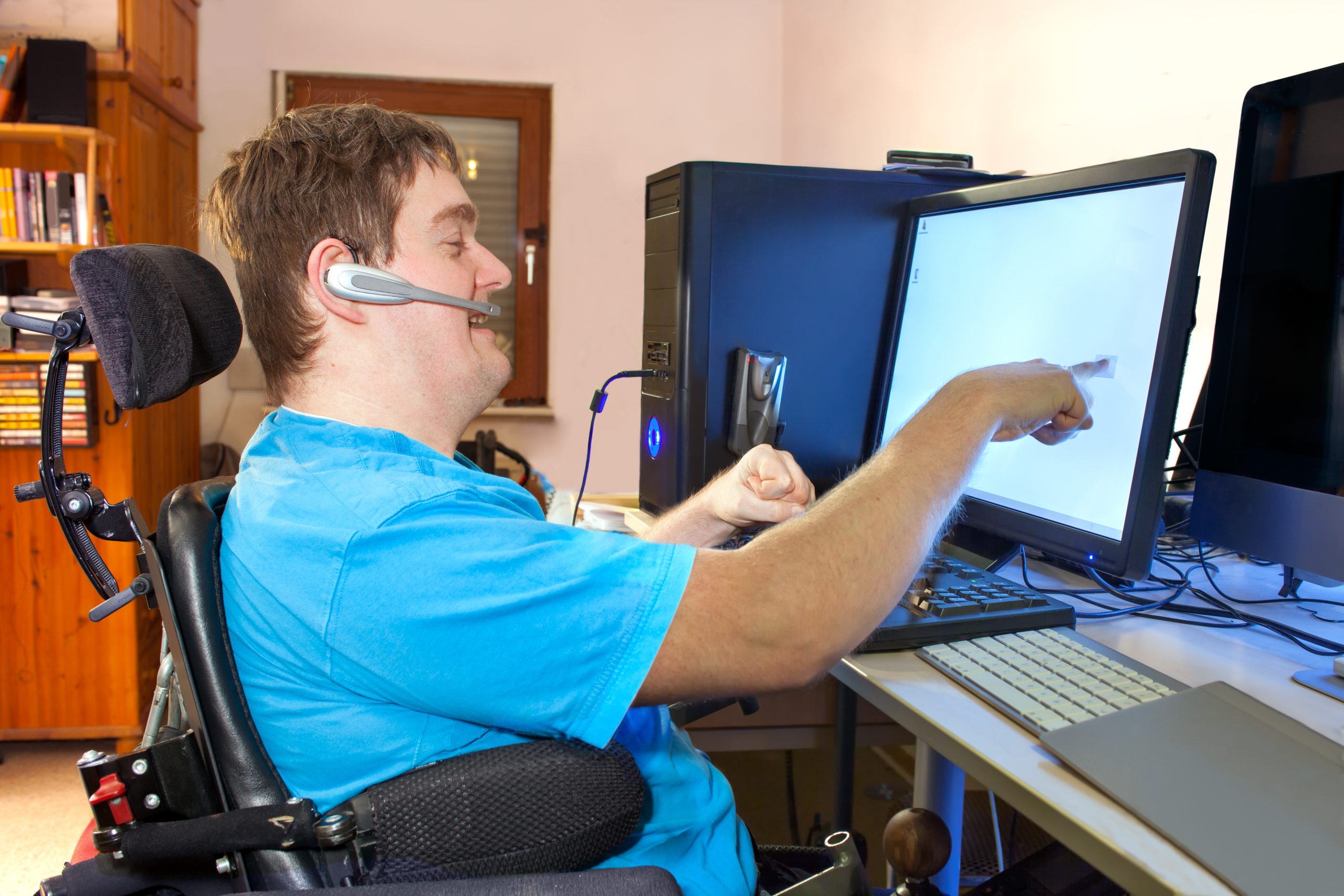 Young man with developmental disabilities sitting in a multifunctional wheelchair, using a computer with a wireless headset, reaching out to touch the touch screen.